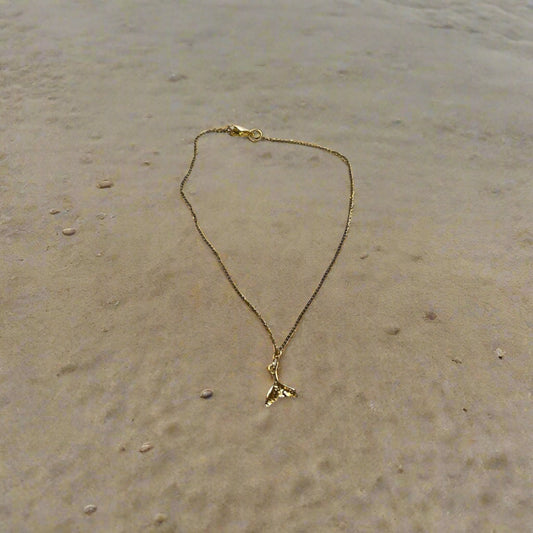 A gold-colored stainless steel anklet in the shape of a whale tail with a small heart-shaped pendant at the end.